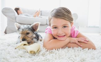 Little Girl Lying On Rug With Yorkshire Terrier Smiling At Camera At Home In The Living Room