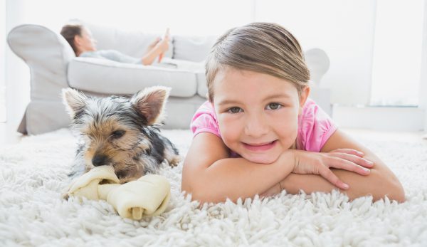 Little Girl Lying On Rug With Yorkshire Terrier Smiling At Camera At Home In The Living Room